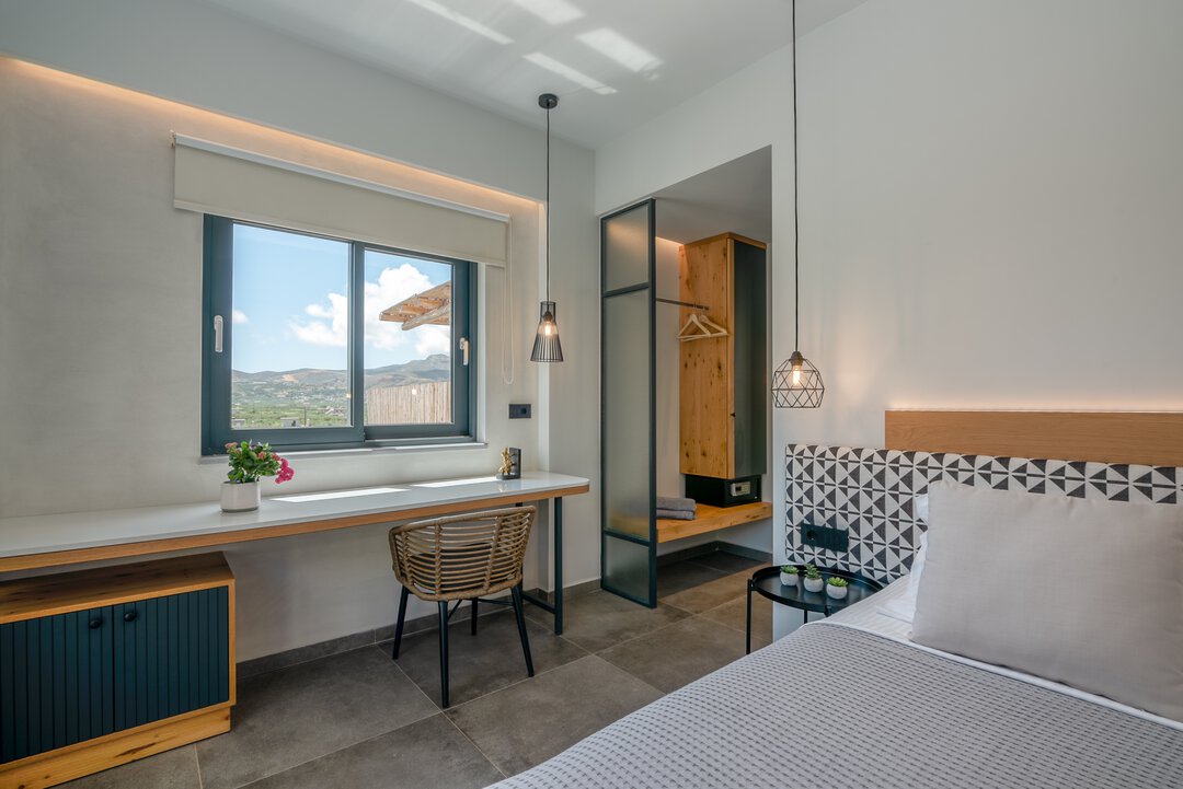 Bright bedroom with workspace desk, rattan chair, geometric pendant lights, and mountain views through window at Nusa Falassarna villa, Crete